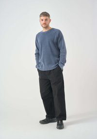 Sudadera de manga larga azul con textura acanalada, corte relajado, combinada con pantalones anchos negros y zapatillas oscuras. Fondo liso.