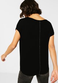 Černý top s krátkým rukávem a volným střihem, s kulatým výstřihem a texturovaným stříbrným švem probíhajícím podél zadní části.