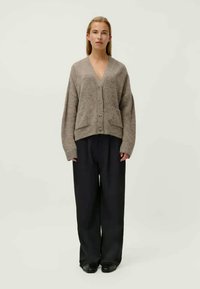 Beige-ylimitoitettu neuletakki V-pääntiellä, napituksella, kahdella etutaskulla ja resorikäänteisillä kalvosimilla, yhdistettynä löysiin mustiin housuihin ja kenkiin.