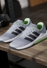 Paire de chaussures de sport grises avec des rayures noires et un intérieur vert, reposant sur une surface en bois près d’un haltère dans une salle de sport.