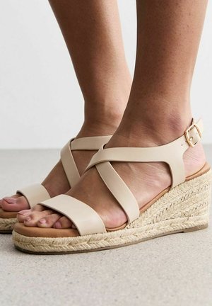 Sandalias de cuña beige con correas cruzadas, hechas de cuero suave. Tacón de cuña envuelto en yute y suela plana. Dedos expuestos visibles.