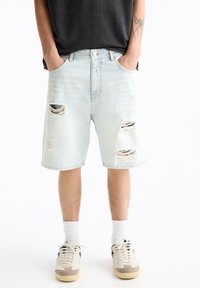 Shorts de mezclilla azul claro con detalles desgastados, que presentan múltiples desgarros grandes y un corte relajado. Completados con un botón de metal y trabillas para cinturón.