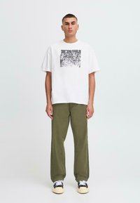 Man standing facing forward wearing a white graphic t-shirt, olive green pants, and black and white sneakers against a plain light background.