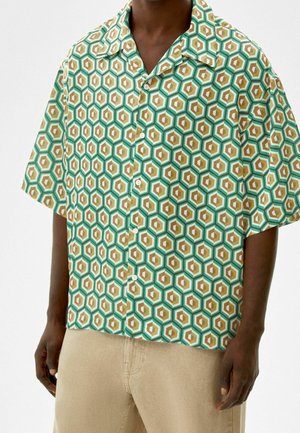 Homme portant une chemise à manches courtes à boutons avec un motif hexagonal vert, beige et blanc, assortie à un pantalon beige clair.