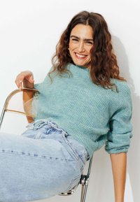 Pull en tricot bleu clair avec une coupe décontractée et des poignets retroussés, associé à un jean en denim clair taille haute, assis sur une chaise en chrome.