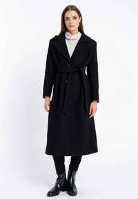 Długi czarny wełniany płaszcz z paskiem w talii, dwoma frontowymi guzikami, szerokimi klapami i długimi rękawami. Noszony na jasnoszarym golfie i czarnych spodniach.