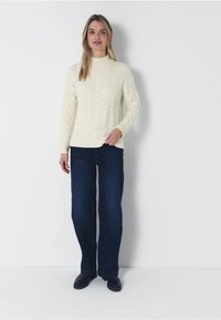 Kabelstickad krämfärgad tröja med hög krage och ribbade manschetter, kombinerad med mörkblå vida jeans och svarta skor, mot en enfärgad bakgrund.