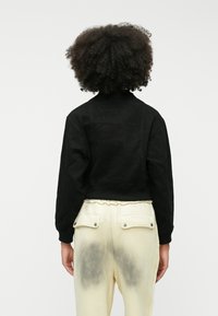 Črna skrajšana jakna z teksturiranim zaključkom, z visokim ovratnikom in rebrastimi manšetami, kombinirana z kremnimi hlačami, ki prikazujejo temne oznake.