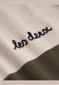 Kötött pulóver, amelynek krémszínű felső része és olajzöld alsó része van, fekete, kurzív betűtípussal hímzett "les deux" felirattal.
