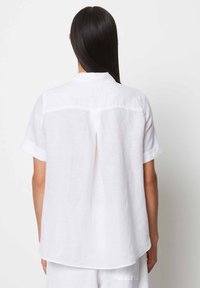 Camicia bianca in lino a maniche corte con colletto classico, dettaglio della plettatura sul retro e vestibilità rilassata. Tessuto liscio e orlo curvo.