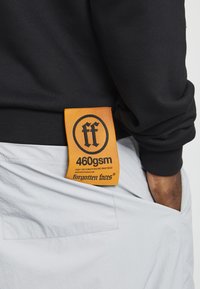 Fekete pulóver, textúrált felülettel, élénk narancssárga címkével, amelyen a "460gsm" felirat és a "forgotten faves" márkaneve található.
