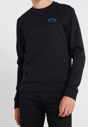 Man draagt een zwarte crewneck trui met een klein blauw "BOSS HUGO BOSS" logo op de linkerborst, gecombineerd met een zwarte broek.