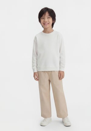 Uśmiechnięte dziecko o czarnych włosach, ubrane w białą bluzę, beżowe spodnie z szerokimi nogawkami i białe trampki, stojące na tle jednolitego tła.