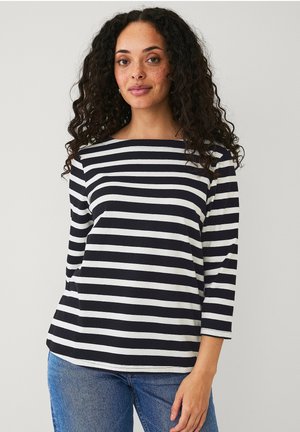 Top a righe bianche e nere con maniche lunghe e scollo a barca, caratterizzato da una vestibilità comoda e materiale in cotone morbido. Abbinato a jeans blu.
