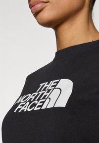 Μαύρο βαμβακερό μπλουζάκι με μεγάλο λευκό λογότυπο «THE NORTH FACE» σε έντονη γραφή στο στήθος. Κοντινή άποψη του λαιμού και του ώμου.