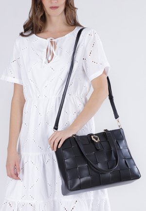 Donna in abito bianco con ricami a traforo che tiene una grande borsa nera intrecciata in pelle con tracolla e dettaglio fibbia.