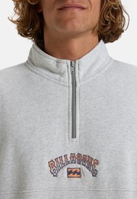Šedý fleecový pulovr se zipovým límcem, s multicolor logem "Billabong" vyšívaným na hrudi. Textura se zdá být měkká.