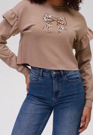 Beiger Langarmpullover mit Rüschen und Leopardenmuster-Schleifendesign, kombiniert mit hoch taillierten blauen Jeans an einer stehenden Person.