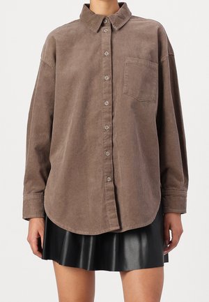 Persoon draagt een bruine corduroy blouse met knopen, een borstzak en lange mouwen, gecombineerd met een zwarte leren geplooide rok.