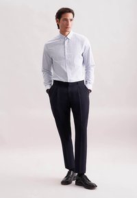 Lichtblauw geruit overhemd, dichtgeknoopt, met zachte kraag. Donkere op maat gemaakte broek, platte voorkant, enkele plooien. Zwarte leren schoenen, klassieke stijl.