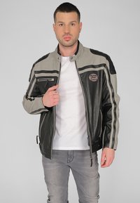 Svart och grå lädermotorcykelfodral med dragkedjefickor, kontrasterande ränder och en rund logotyp-patch på bröstet. Mjukt snitt.