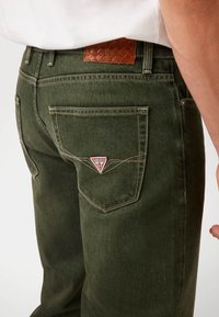 Tamsiai žali džinsai su trikampiu raudonu ir baltu pleistru ant galinio kišenės bei tekstūruotu ruduo odiniu diržu.