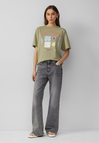 Olijfgroene t-shirt met een grafische print in rood, blauw en geel, gecombineerd met hoogopgetrokken, wijde grijze jeans en witte sandalen.