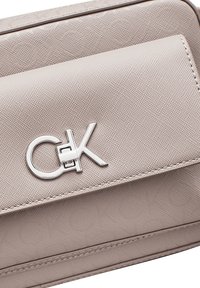 Beige texturovaná kabelka s přední klapkou, která má stříbrný logo detail. Materiál se zdá být syntetický s hladkým povrchem.