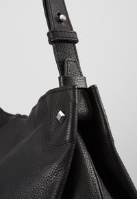 Svart texturerat läderväska med en stor öppning, med detaljer av silverfyrkantiga nitar och justerbar rem med tryckknapp.