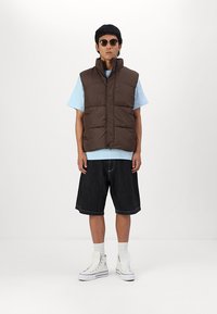 Brown puffer vest, sleeveless with vertical stitching, paired with a light blue t-shirt and dark denim shorts. White high-top sneakers worn.