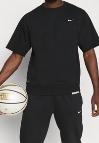 Hombre con ropa deportiva negra de Nike sosteniendo un balón de baloncesto blanco y dorado de Nike sobre un fondo gris liso.