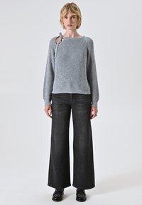 Donna che indossa un maglione grigio a maglia con dettaglio di legatura sulle spalle, jeans neri a gamba larga e scarpe slip-on nere con borchie, in piedi contro uno sfondo bianco.