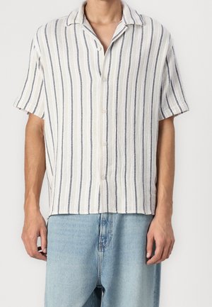 Person trägt ein weißes kurzärmeliges Hemd mit schwarzen vertikalen Streifen und hellblauen Jeans aus Denim und steht vor einem schlichten Hintergrund.