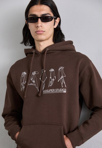 Brown hoodie featuring white graphic prints of figures, drawstring hood, and front pocket detail. Made from a soft material.