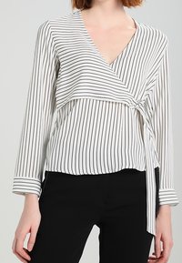 Weiße Bluse mit schwarzen vertikalen Streifen, Wickel-Design, langen Ärmeln, V-Ausschnitt und einem gebundenen Detail an der Taille. Glatte Textur.