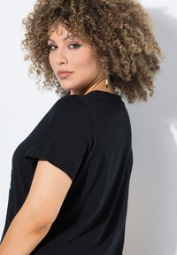Černé tričko s krátkým rukávem a kulatým výstřihem. Viditelné kudrnaté vlasy a kruhové náušnice, které ukazují přirozený tón pleti a jemnou strukturu.