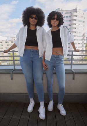 Duas pessoas a usar tops cropped pretos e casacos brancos estão lado a lado. Elas usam jeans skinny azuis e ténis brancos. Um acessório de corrente adorna as suas cinturas.