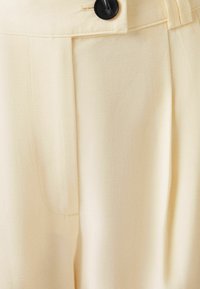 Lichtbeige broeken van een gladde stof, met een enkele zwart knoopsluiting, plooien en zijzakken.