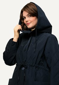 Vadderad, marinblå jacka med huva, snörning och knäppning. Den har en insydd midja och två framfickor. Slät textur.