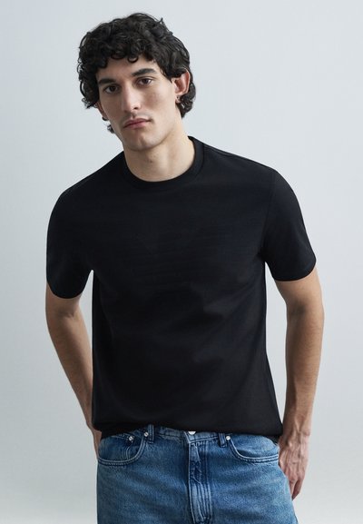 Sort bomulds t-shirt med et diskret præget logo på brystet. Korte ærmer, rund halsudskæring og en glat tekstur. Kombineret med blå jeans.