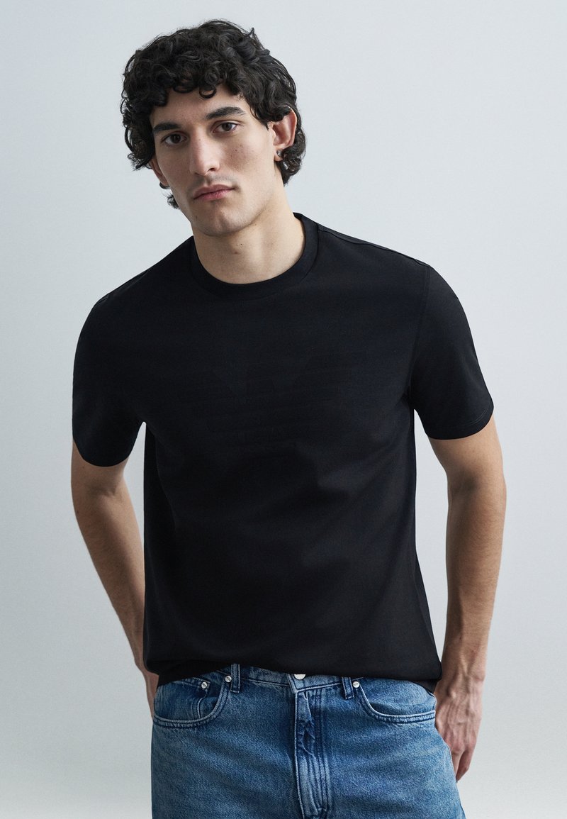 Μαύρο βαμβακερό T-shirt με διακριτικό ανάγλυφο λογό�τυπο στο στήθος. Κοντά μανίκια, στρογγυλή λαιμόκοψη και απαλή υφή. Συνδυασμένο με μπλε τζιν.