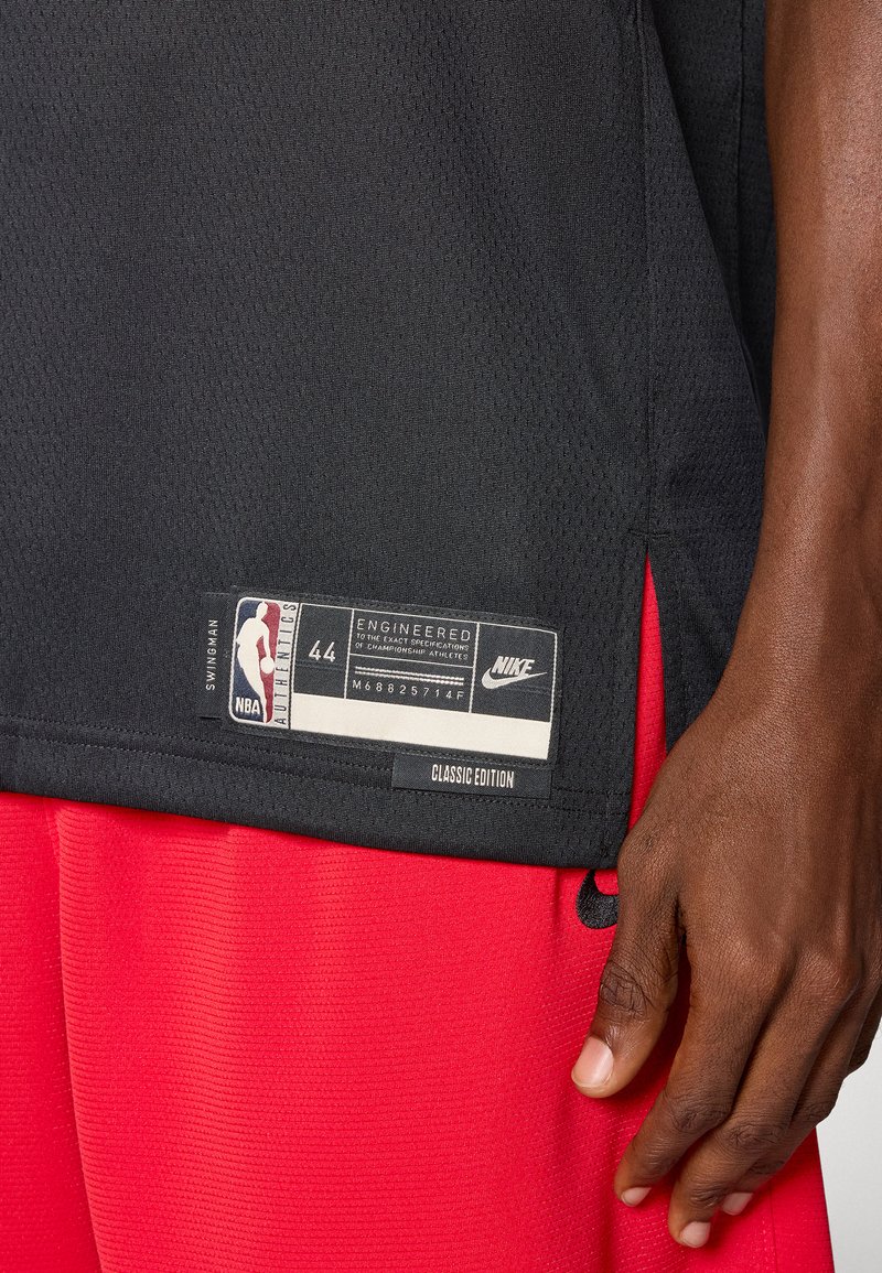 Black basketball jersey with mesh texture, tag at hem displaying size 44, NBA logo, and Nike branding; red shorts partially visible.