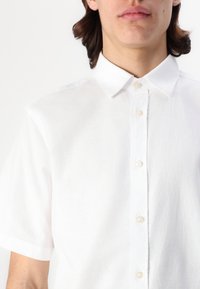 Camicia bianca a maniche corte realizzata in tessuto leggero, con colletto a punta e chiusura frontale con bottoni di colore chiaro. Texture liscia.
