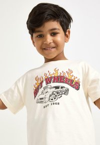 Valkoinen puuvillapaita, jossa on punainen "Hot Wheels" -grafiikka, jossa on liekkejä, auton kuvastoja ja teksti "EST 1968". Lyhyet hihat, normaali istuvuus.