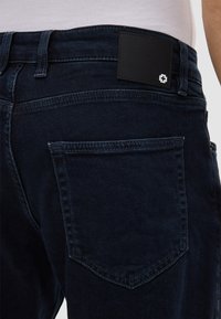Jeans in denim blu scuro con una toppa in pelle nella parte posteriore della vita, dotati di due tasche posteriori e dettagli di cucitura sottili.