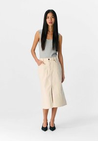 Gonna di jeans beige chiaro con uno spacco frontale e tasche, abbinata a un top senza maniche grigio. La modella indossa scarpe con tacco a punta nere.