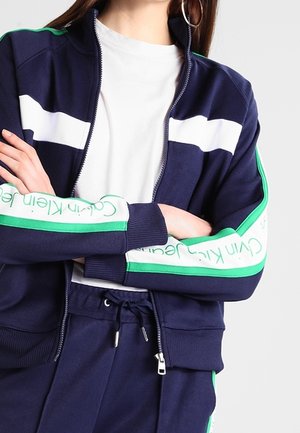 Osoba ubrana w granatową kurtkę zapinaną na zamek oraz pasujące spodnie z białymi i zielonymi paskami oraz napisem „Calvin Klein Jeans” na rękawach.