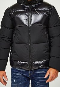 Svart puffjacka med en glansig övre del, dragkedjestängning och sidofickor. Den har en strukturerad kontrasterande nedre del och en huva.