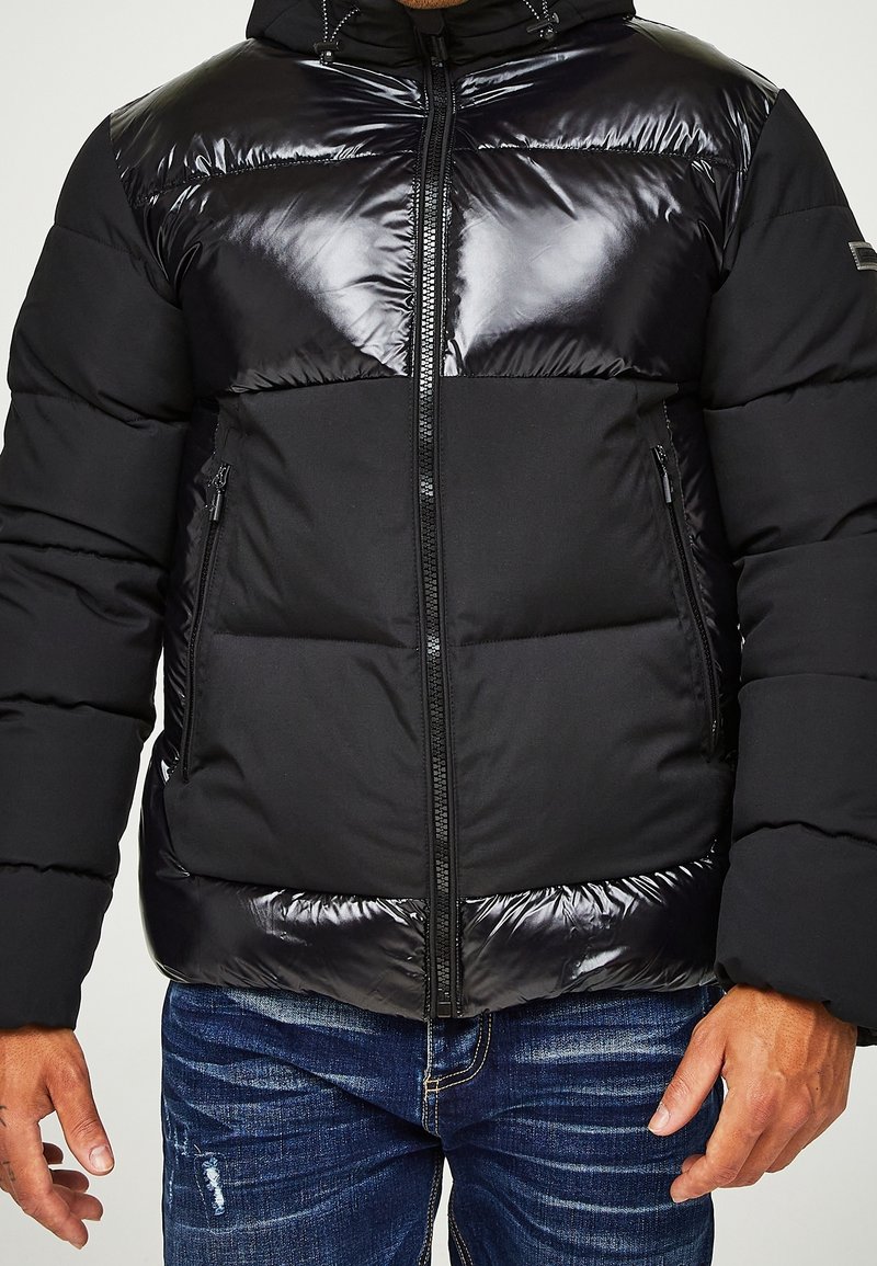 Svart puffjacka med en glansig övre del, dragkedjestängning och sidofickor. Den har en strukturerad kontrasterande nedre del och en huva.