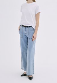 Valkoinen puuvillapaita lyhyillä hihoilla, yhdistettynä löysiin vaaleansinisiin farkkuihin, joissa on sivutaskut ja musta vyö. Mustat kärkitossut.